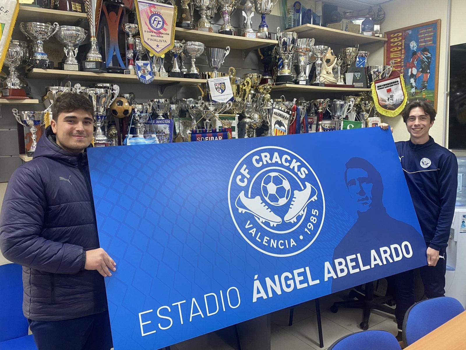 Dos jóvenes sostienen el cartel azul del Estadio “Ángel Abelardo” del CF CRACKS, en la sala de trofeos